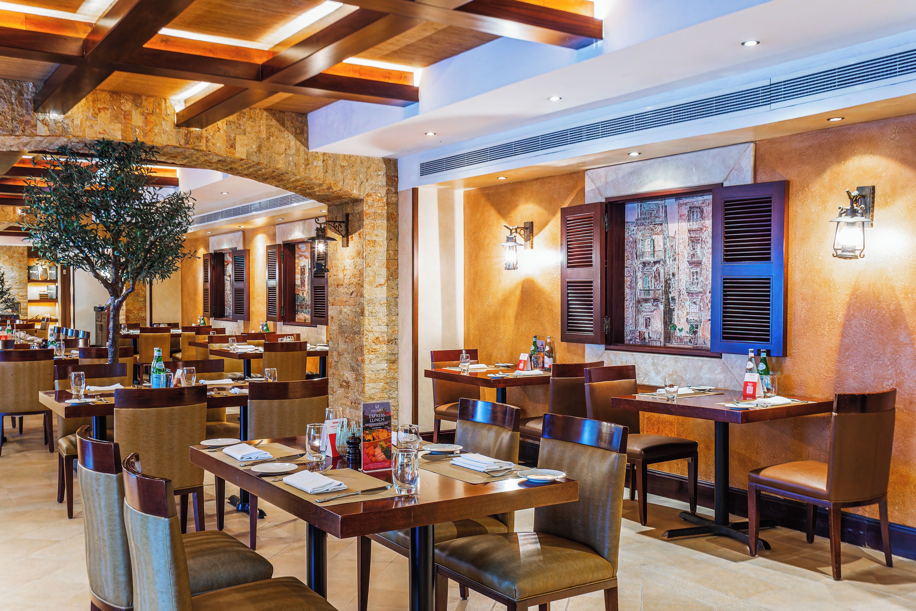 A restaurant with multiple tables and chairs, wooden beams on the ceiling, and a large tree in the center.