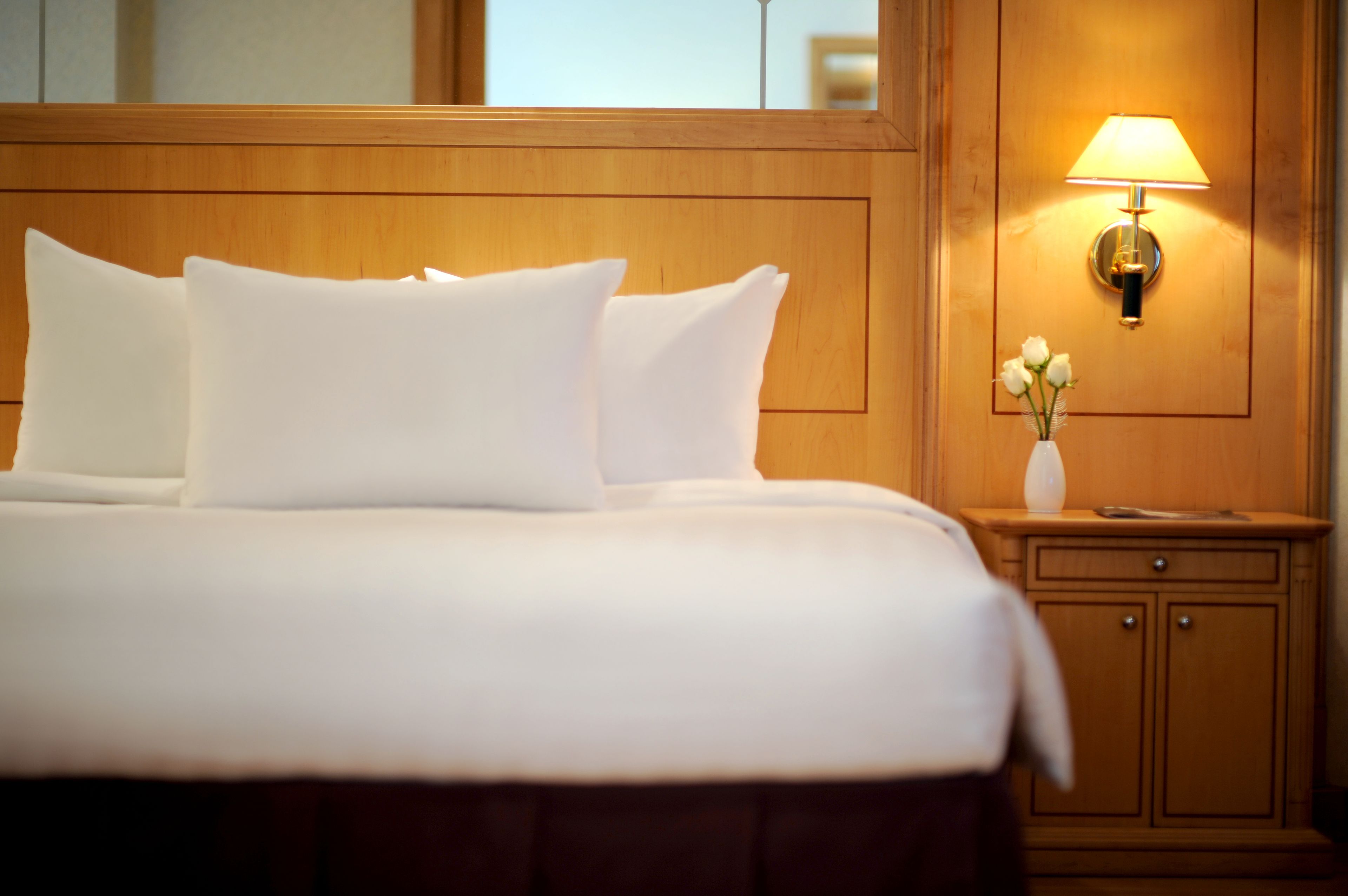 Een net opgemaakt bed met witte lakens en kussens, een nachtkastje met een lamp en een bloemenvaas, en houten lambrisering aan de muur.