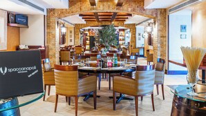 Un restaurante con un arco de piedra, vigas de madera en el techo y una planta central. Las mesas están dispuestas con sillas, y hay un portátil sobre un soporte.