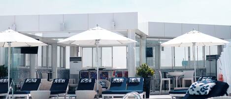 A poolside area with lounge chairs and umbrellas, a glass-enclosed bar area, and a clear view of the sky.