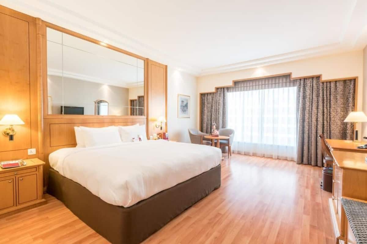 Een hotelkamer met een groot bed, een bureau, een stoel, een klein tafeltje, een spiegel en een raam met gordijnen.