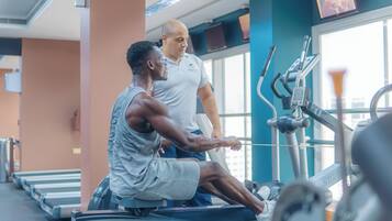 Une salle de sport équipée de matériel d’exercice, un homme utilisant un appareil de rame, et une autre personne qui l’aide.