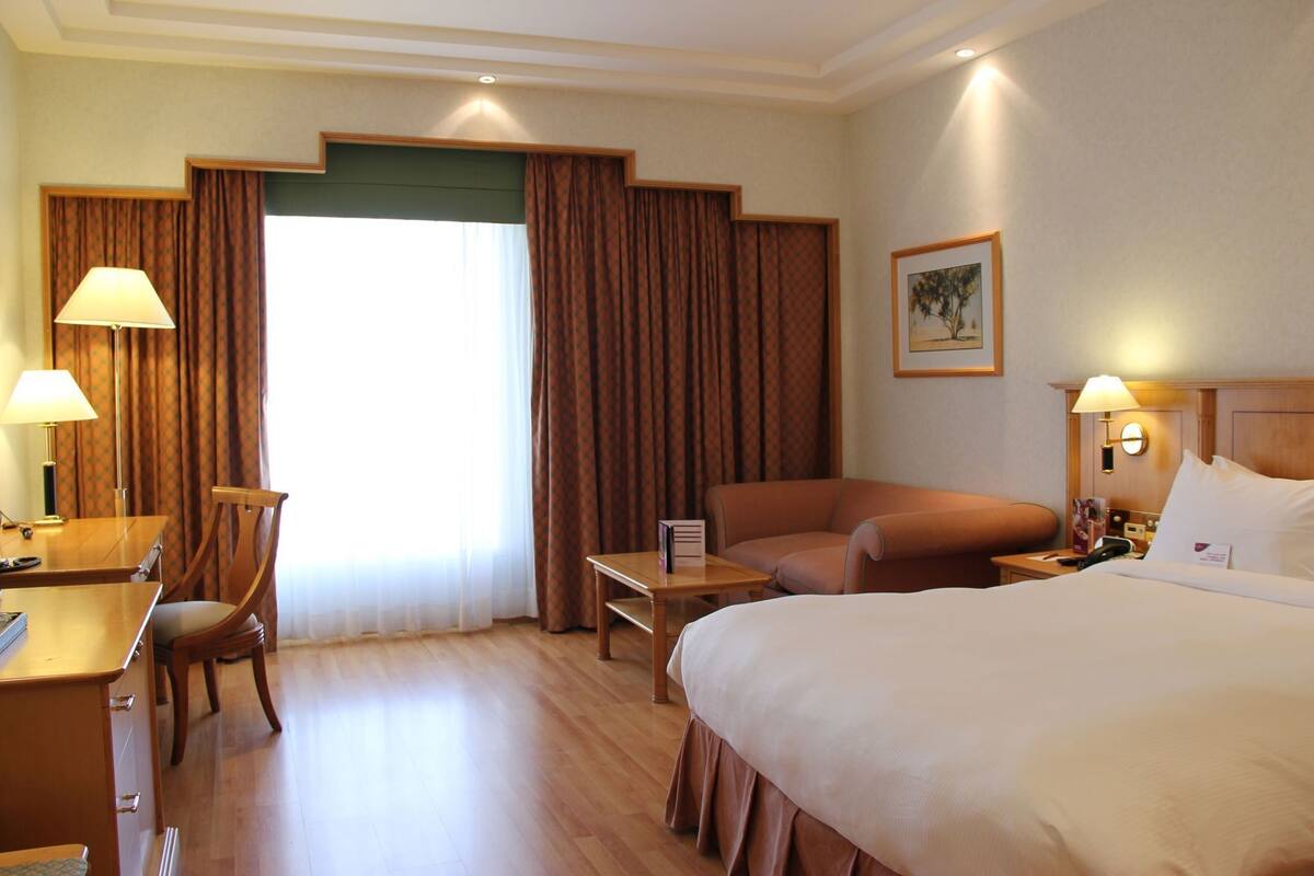 A hotel room with a bed, a desk, a chair, a small table, and a nightstand.