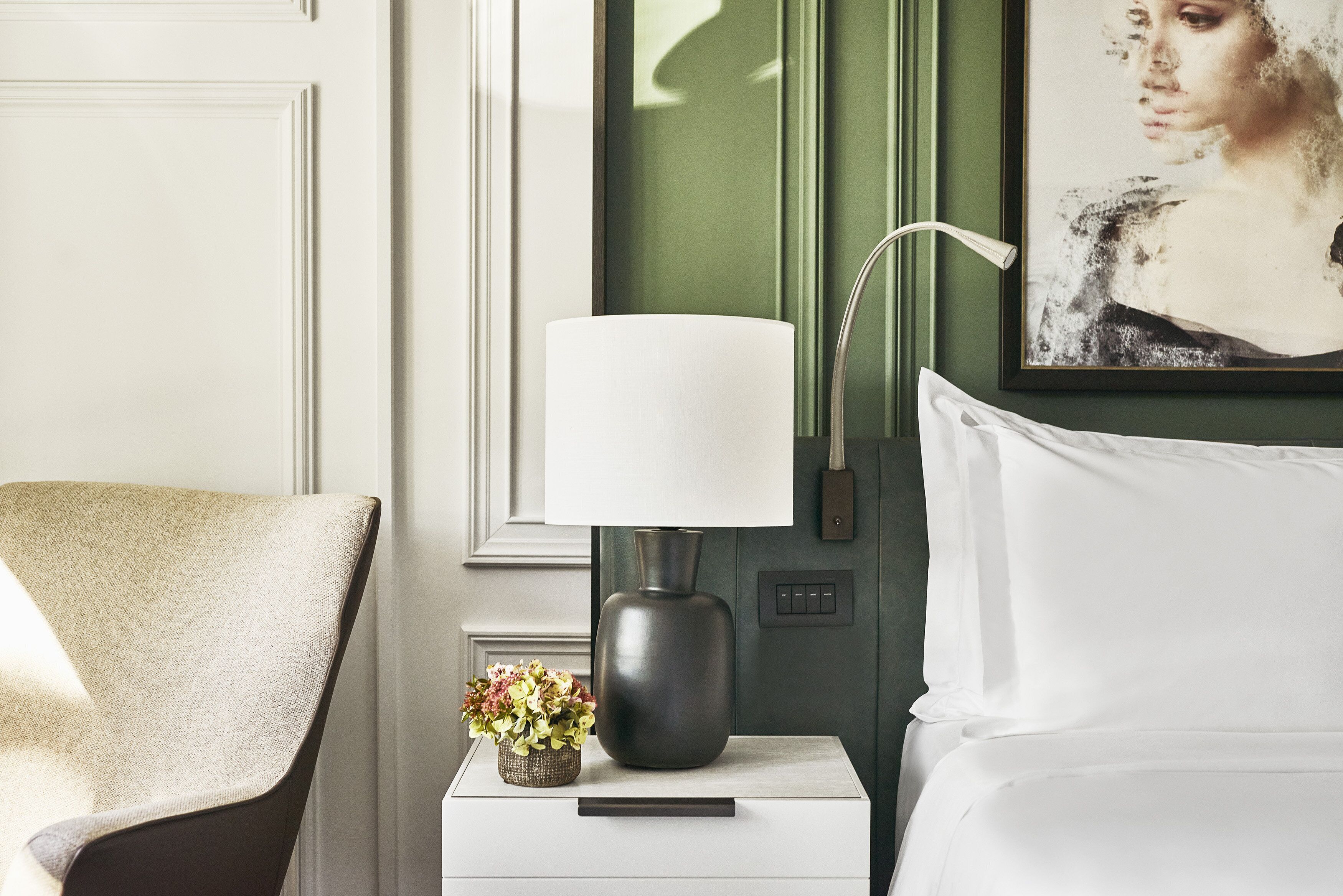 Un dormitorio ordenado con una cama blanca, una mesita de noche con una lámpara, un pequeño jarrón con flores y un cuadro en la pared.