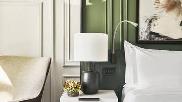 Un dormitorio ordenado con una cama blanca, una mesita de noche con una lámpara, un pequeño jarrón con flores y un cuadro en la pared.