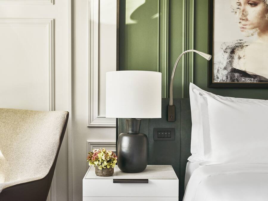 Un dormitorio ordenado con una cama blanca, una mesita de noche con una lámpara, un pequeño jarrón con flores y un cuadro en la pared.