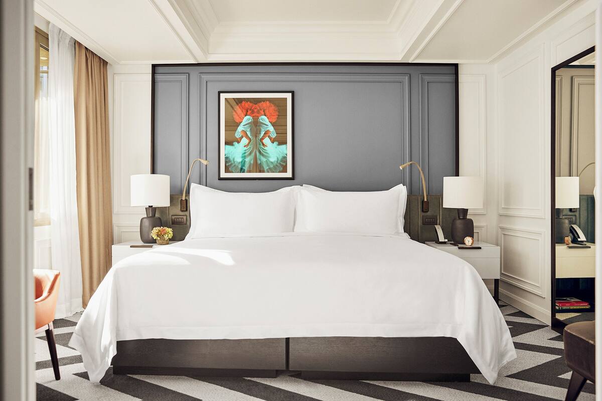 A hotel room with a large bed, two bedside tables with lamps, a framed artwork above the bed, and a view of the hallway.