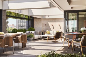 A modern outdoor lounge area with a dining table, sofas, and chairs, featuring a view of the cityscape through large windows.