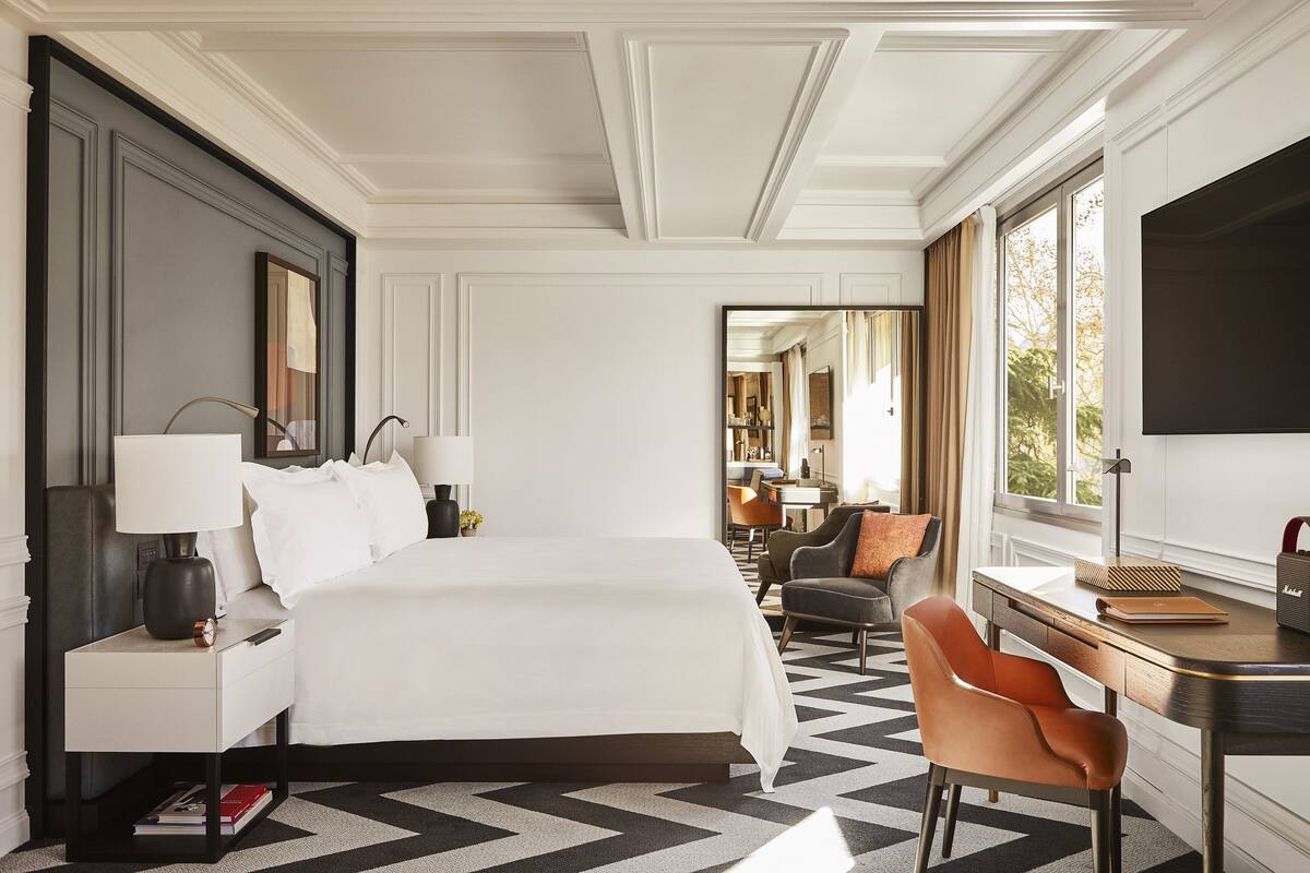 A hotel room with a large bed, a desk with a chair, a nightstand with a lamp, and a large window.