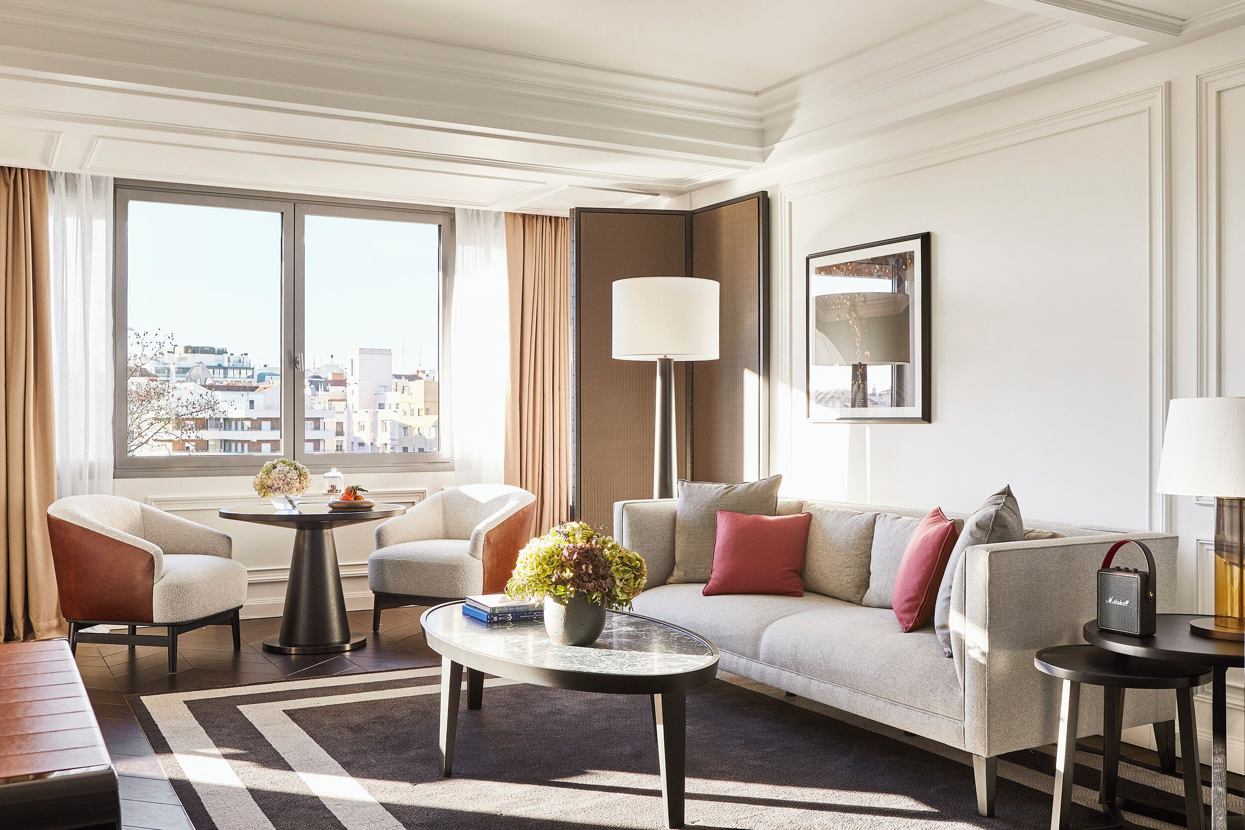 Quarto de hotel moderno com sofá, poltronas, mesa de centro e uma grande janela com vista para a cidade.