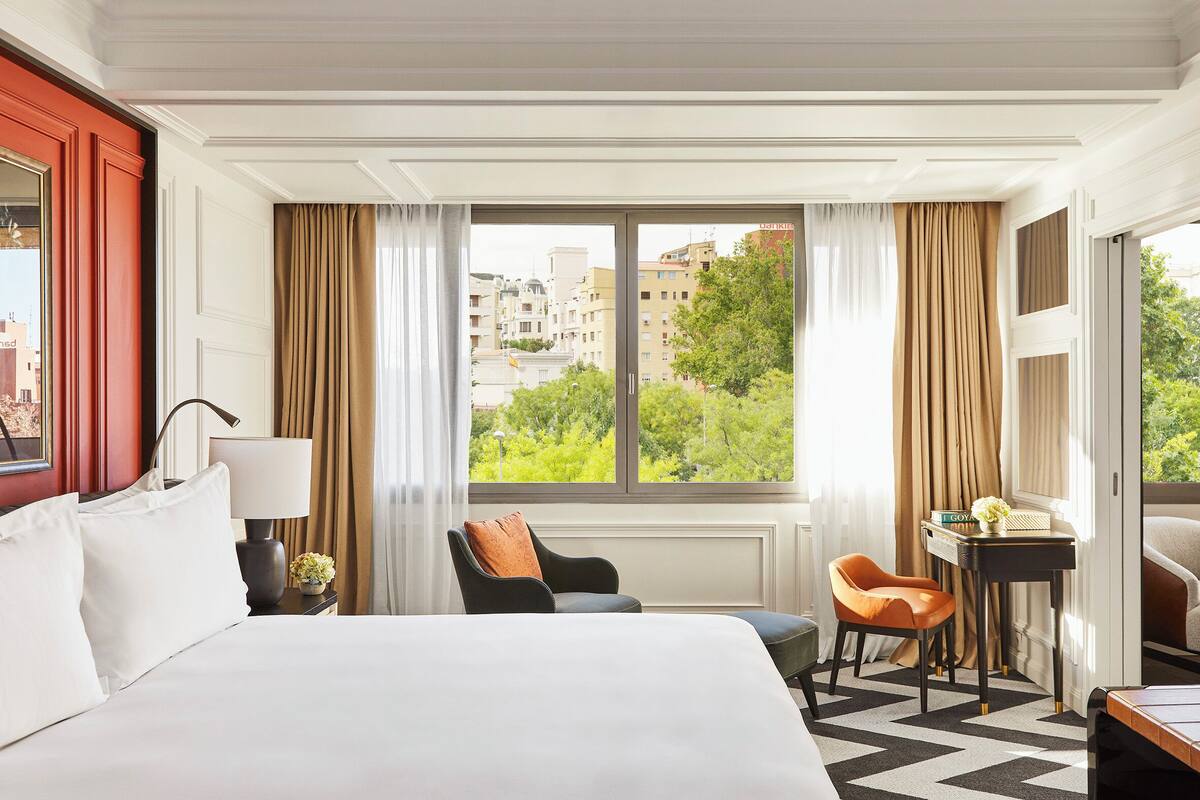 A hotel room with a large bed, a desk, a chair, and a view of buildings and trees.