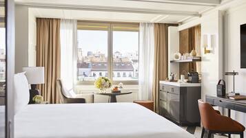 Habitación de hotel con una cama grande, un escritorio, una silla, una mesita con un florero y vistas a edificios a través de la ventana, además de un minibar.
