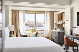 Ein Hotelzimmer mit einem großen Bett, einem Schreibtisch, einem Sessel, einem kleinen Tisch mit Blumen und Blick auf Gebäude durch das Fenster, sowie einer Minibar.