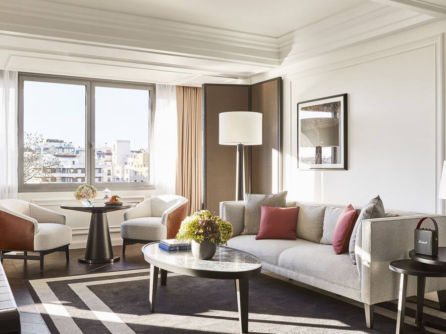 Quarto de hotel moderno com sofá, poltronas, mesa de centro e vista para a cidade através de grandes janelas.