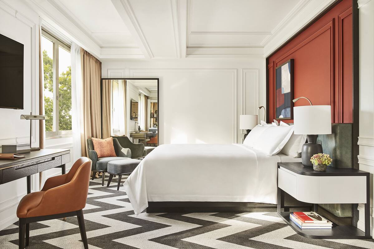 A hotel room with a large bed, a desk with a chair, a TV, and a nightstand with a lamp.