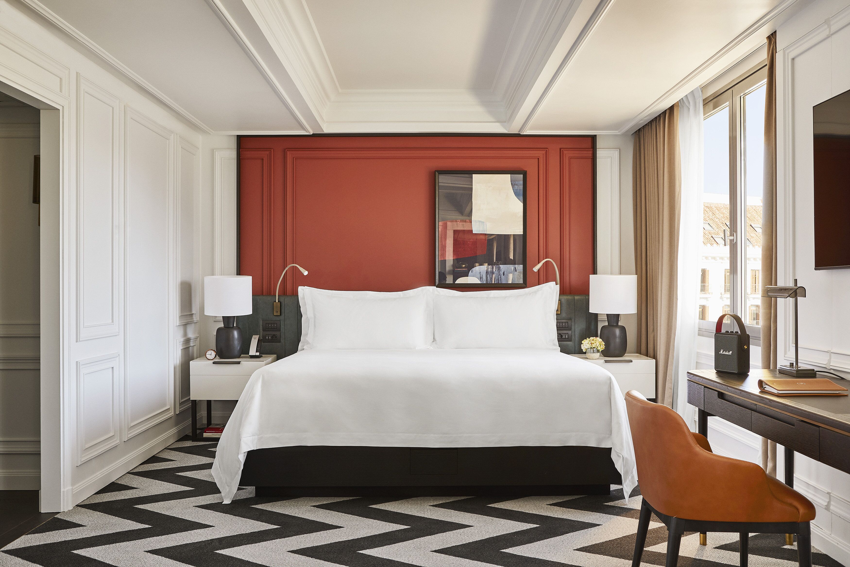 A hotel room with a large bed, two bedside tables with lamps, a desk with a chair, and a large window with curtains.