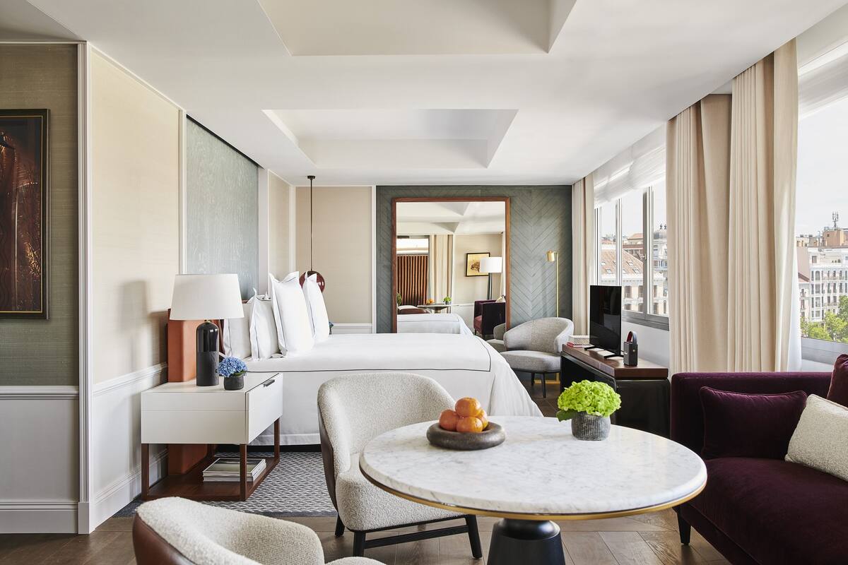 A hotel room with a bed, a desk, a round table with a bowl of fruit, a chair, and a purple sofa.