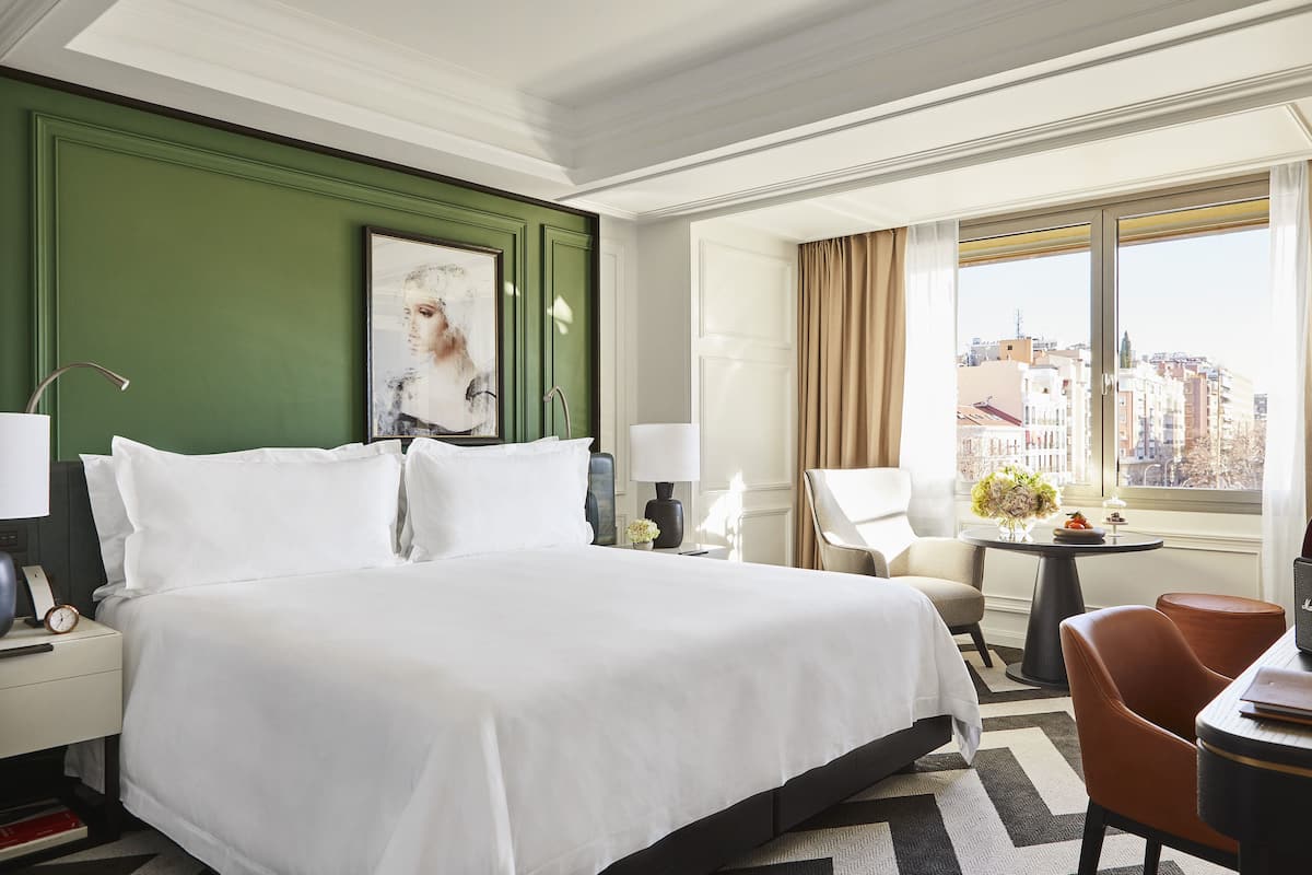 A hotel room with a large bed, a desk with a chair, a small table with flowers, and a view of buildings through the window.