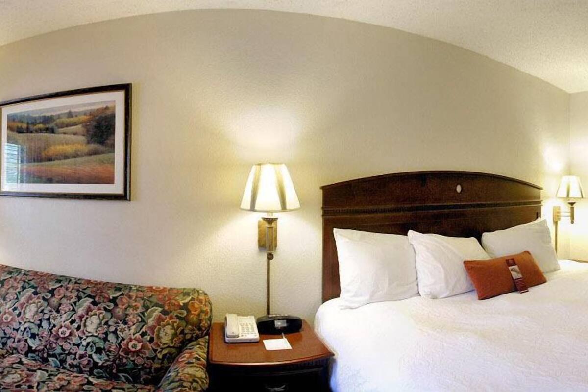 A hotel room with a bed, a floral sofa, a nightstand with a lamp, and a framed picture on the wall.