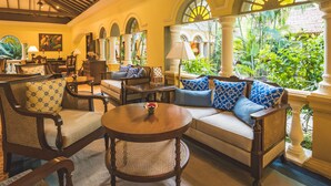 A well-lit, open-plan living area with a sofa set, armchairs, and a coffee table. There are multiple windows with arched tops, allowing natural light to fill the space. - Taj Holiday Village Resort & Spa (Candolim)