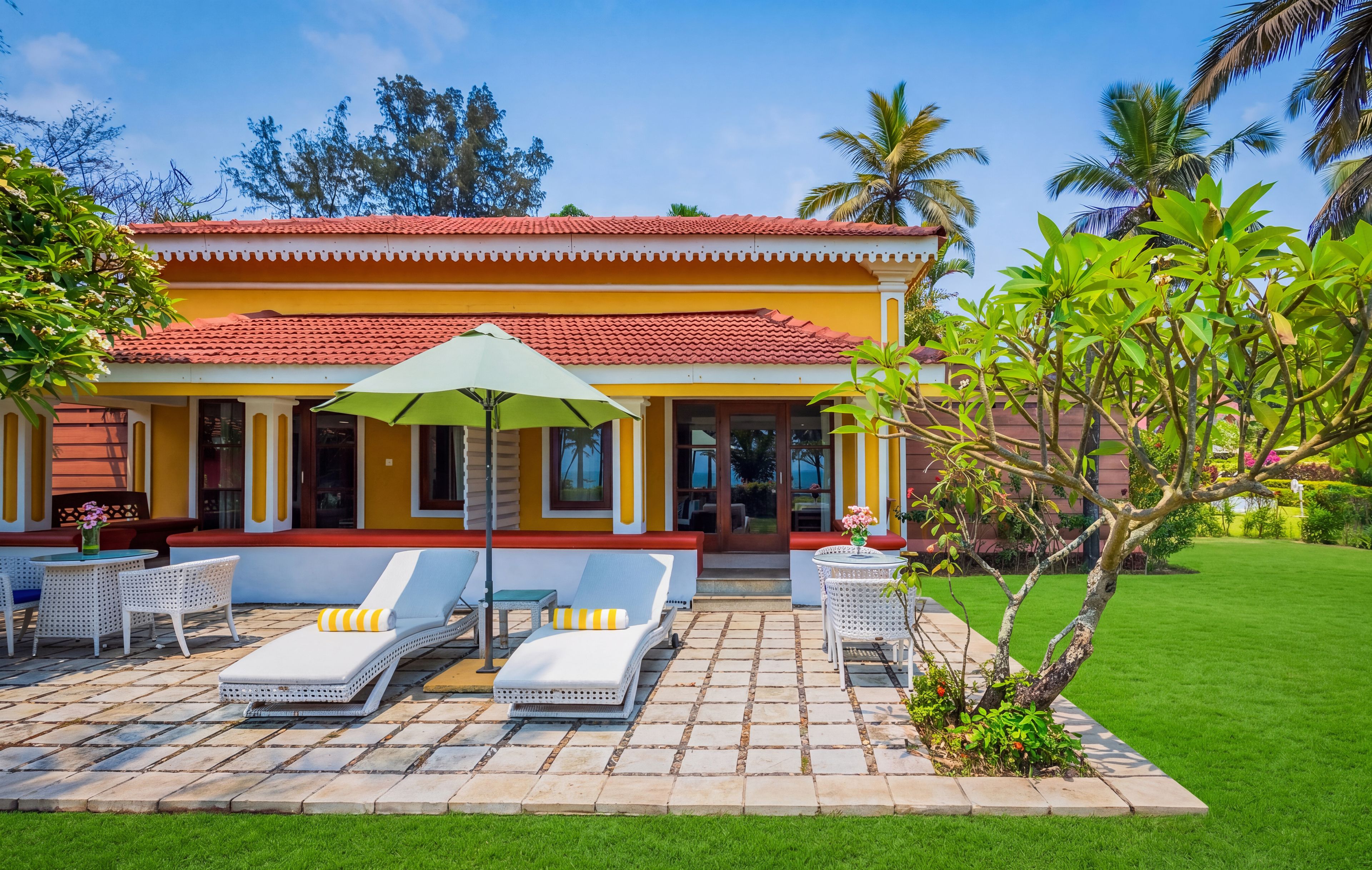 A yellow building with a red roof, outdoor seating with white chairs and a green umbrella, and a well-maintained lawn.