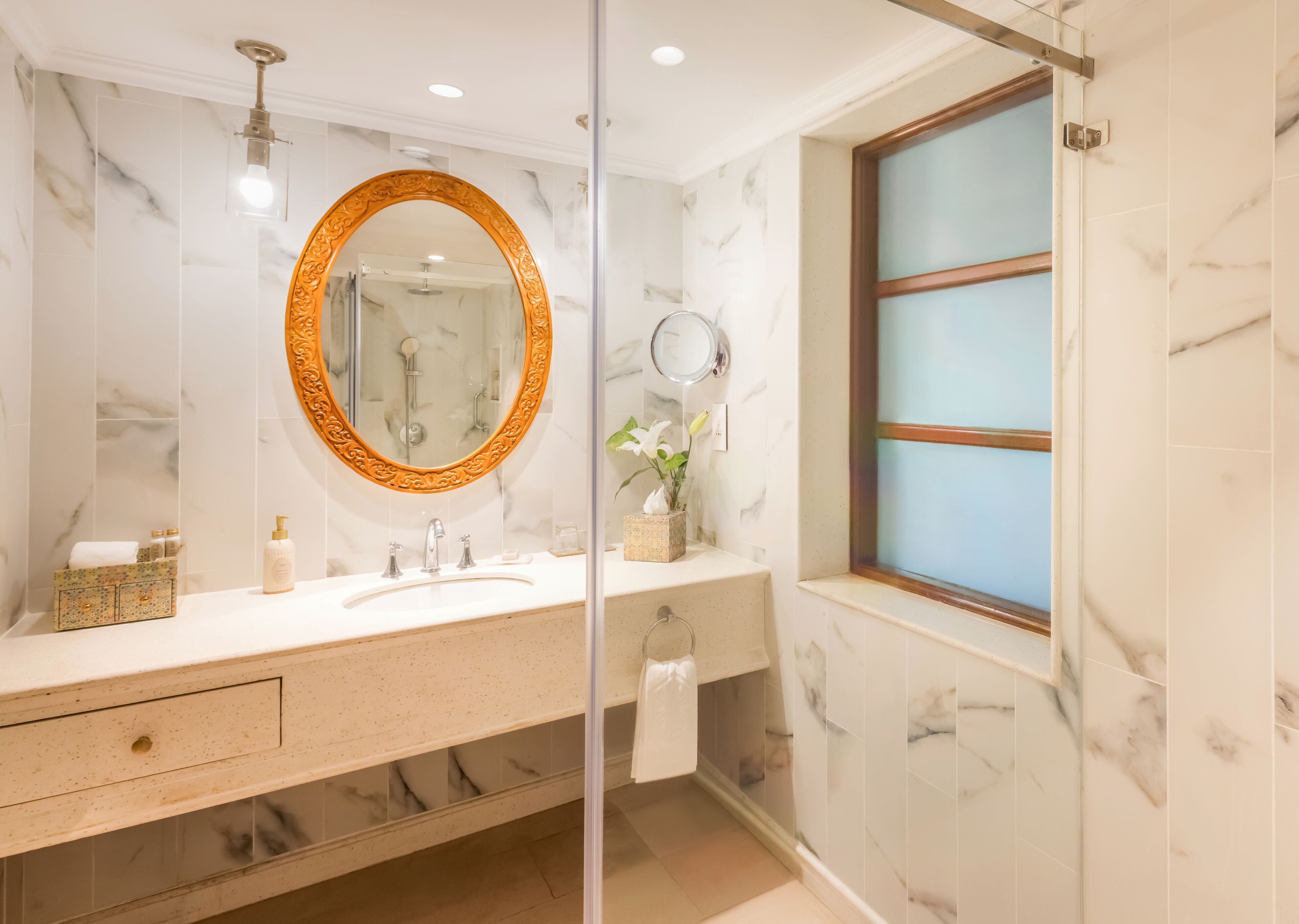 Un baño con pared de mármol, un espejo redondo con marco ornamentado, un lavabo y una ventana con vidrio esmerilado.