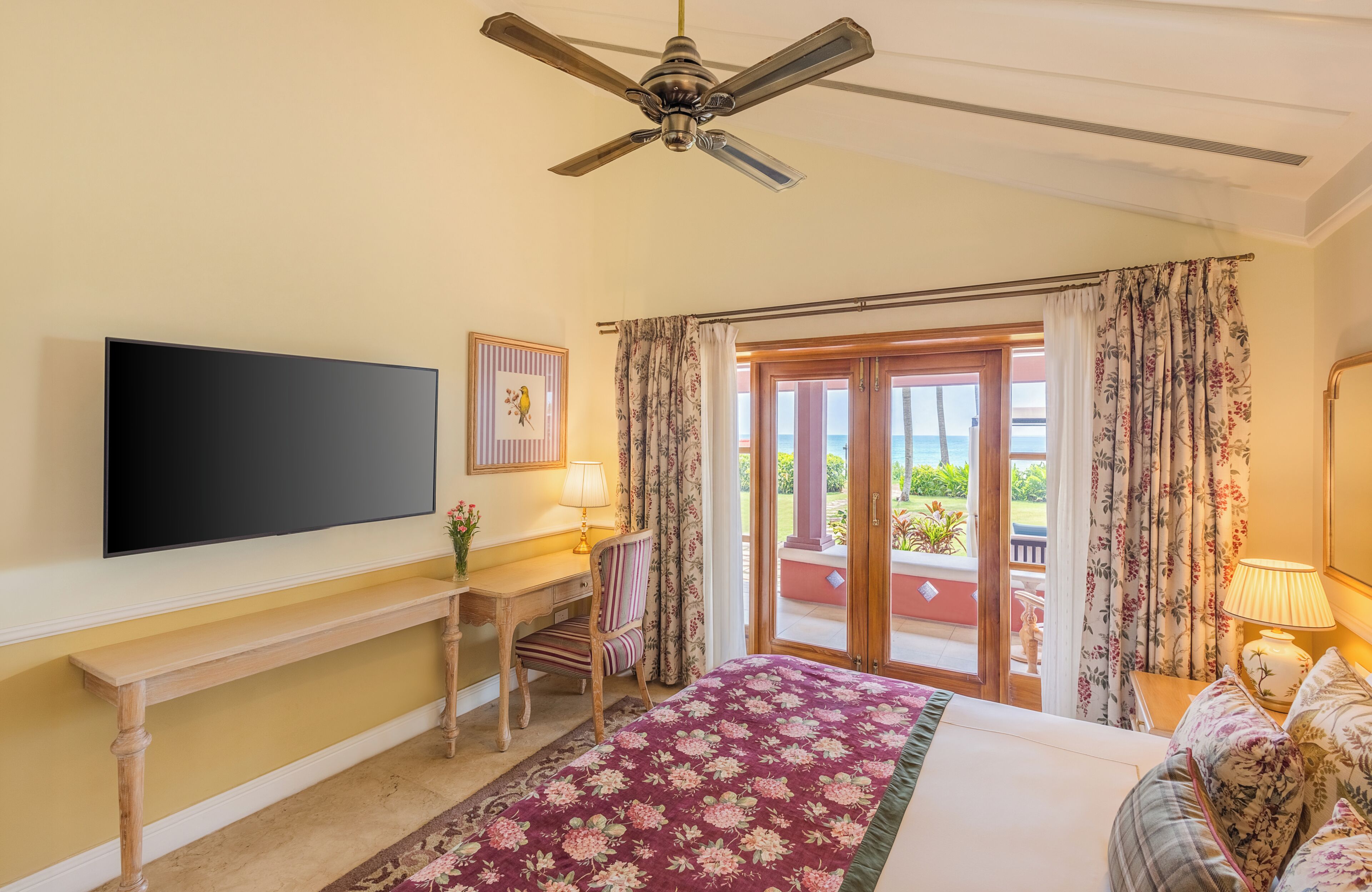 A hotel room with a bed, a desk, a chair, a television, a ceiling fan, and a view of the outdoors.