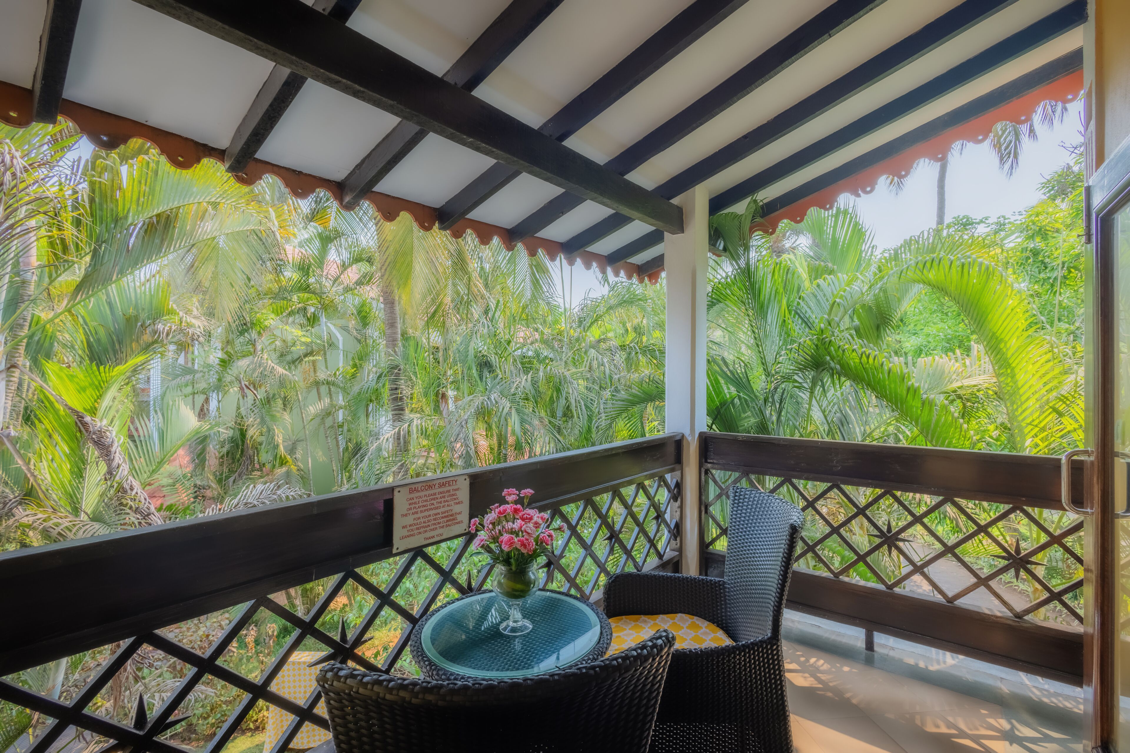 Un balcón con vistas a vegetación exuberante, una silla de mimbre, una mesa de vidrio con un florero y un letrero de seguridad.