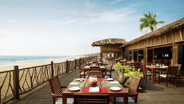 A beachside restaurant with wooden tables and chairs, a thatched roof, and a view of the ocean. - Taj Holiday Village Resort & Spa (Candolim)