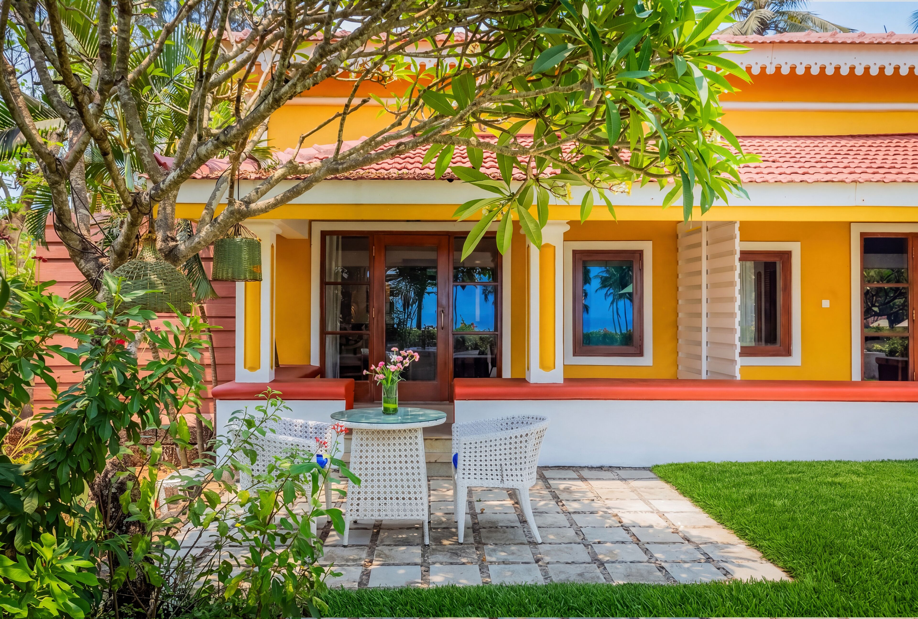 A yellow building with a red-tiled roof, a glass door entrance, and a small patio with a table and chairs.