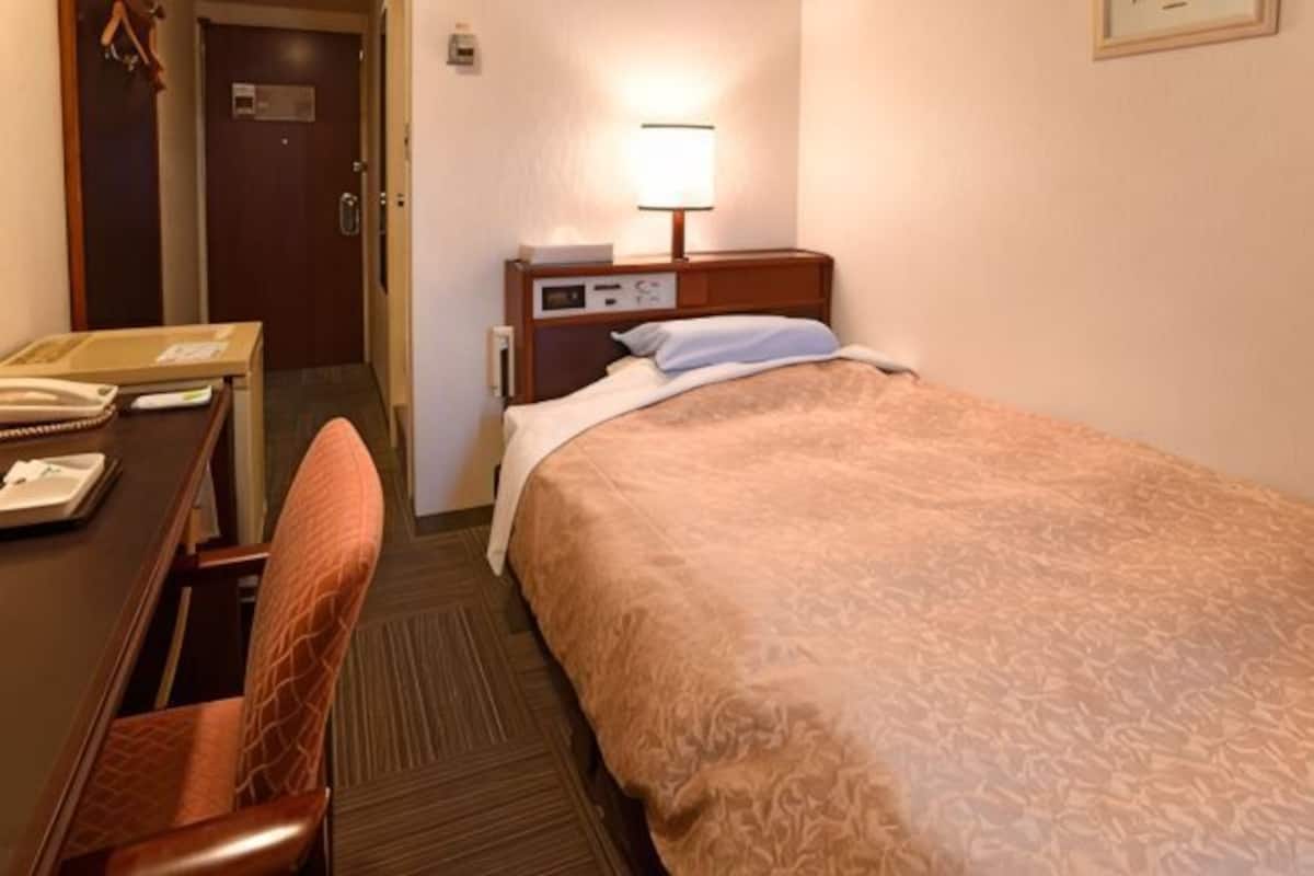 A hotel room with a bed, a desk, a chair, a lamp, and a door.