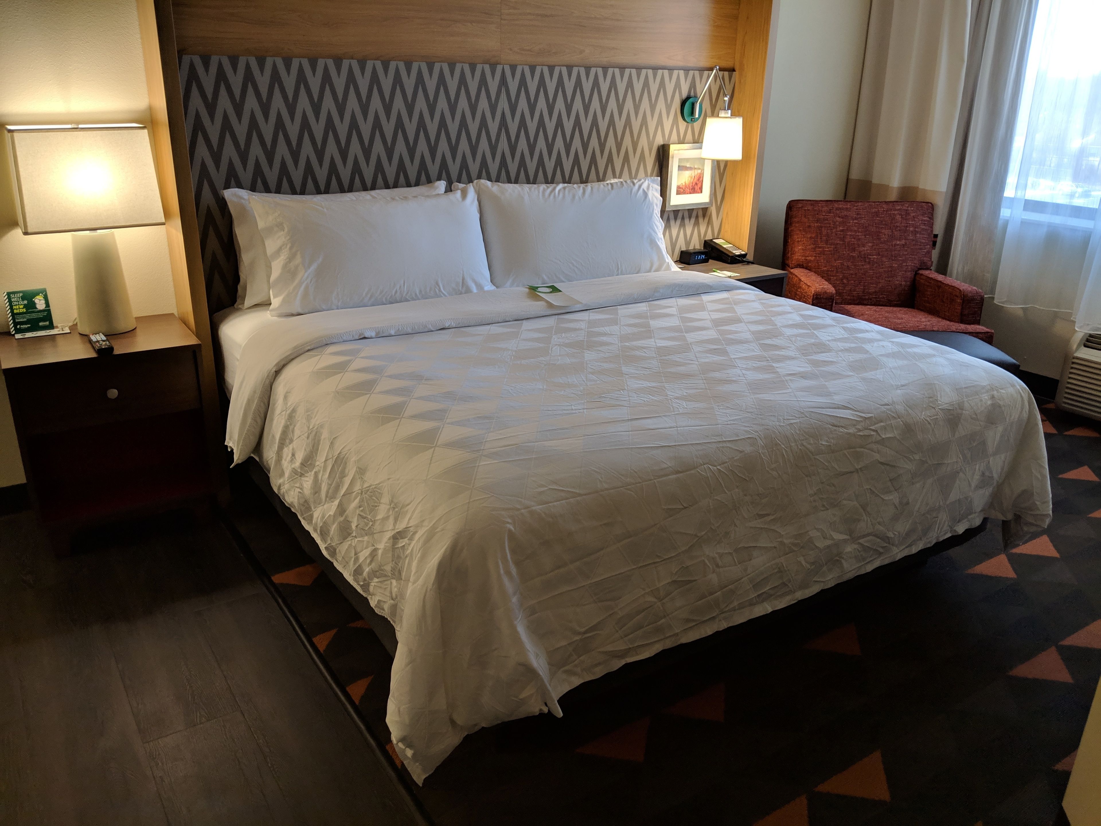 Una habitación de hotel con una cama grande, un cabecero de madera, una mesita de noche con una lámpara, un sillón rojo y vistas al exterior por la ventana.