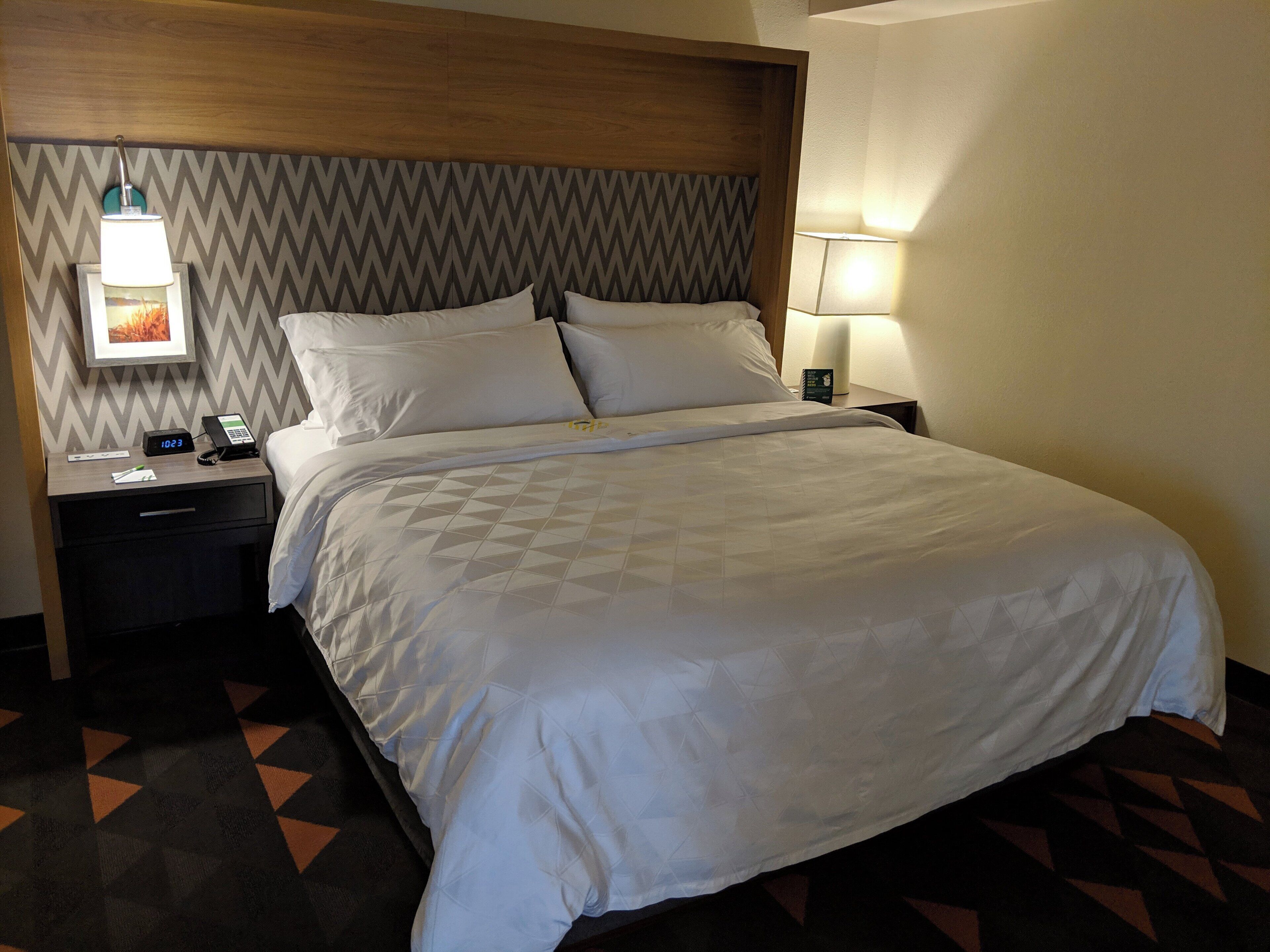 A hotel room with a bed, a nightstand, a lamp, and a wall clock.