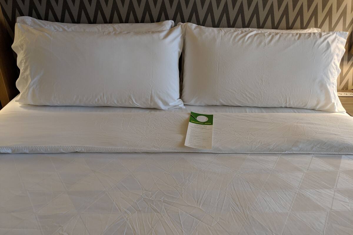 A neatly made bed with white linens and pillows, a green breakfast menu tag, and a patterned wall in the background.