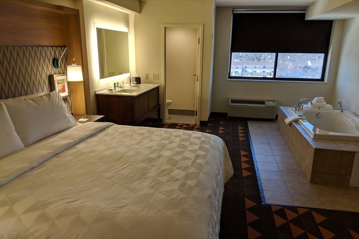 A hotel room with a large bed, a bathroom with a bathtub, a sink, and a window with a view of buildings.