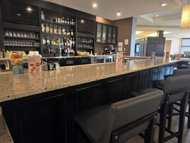 Una zona de bar con una barra larga, taburetes, estantes con botellas y un televisor.