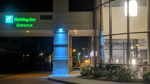 Ingången till Holiday Inn på natten, med upplyst skyltning, en glasfasad och ett anlagt område med växter.