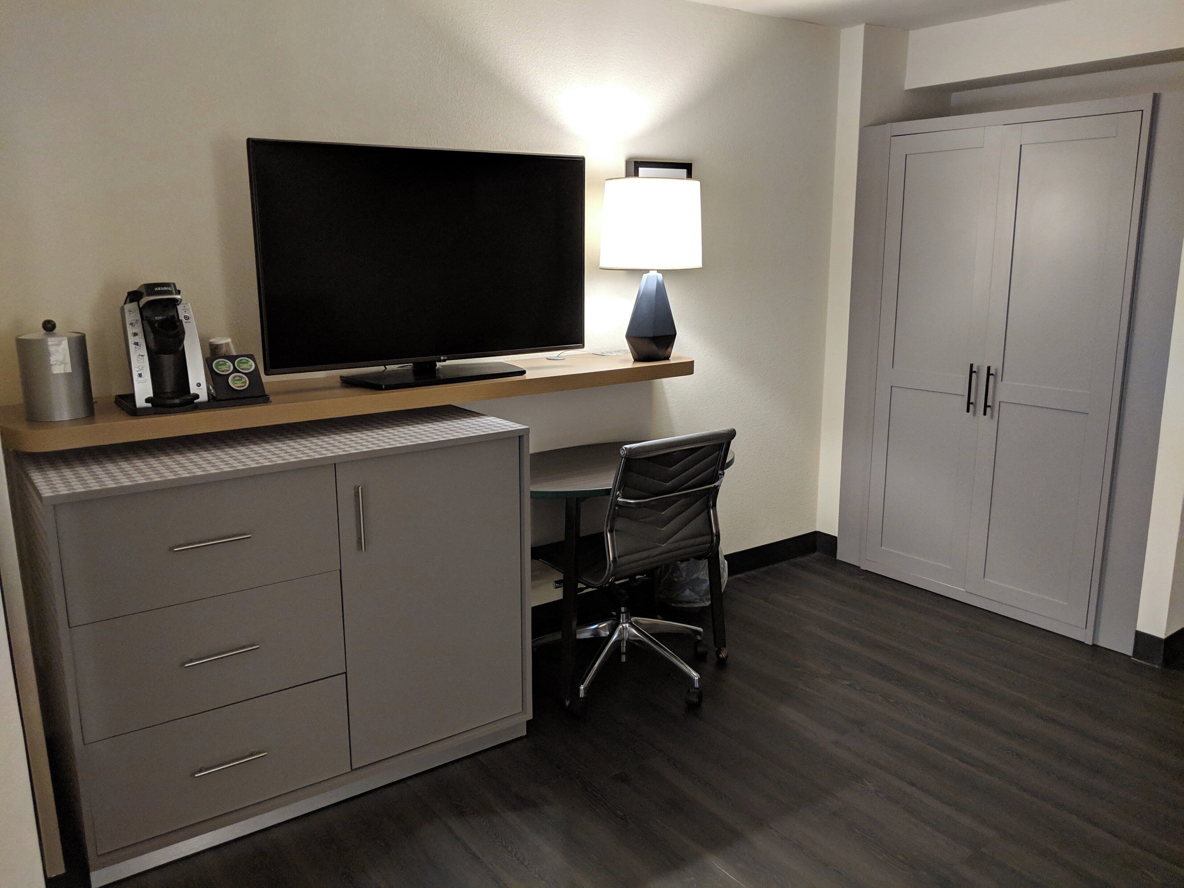 A hotel room with a flat-screen TV, a desk with a lamp, a chair, a filing cabinet, and a wardrobe.
