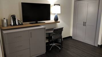 A hotel room with a flat-screen TV, a desk with a lamp, a chair, a filing cabinet, and a wardrobe.