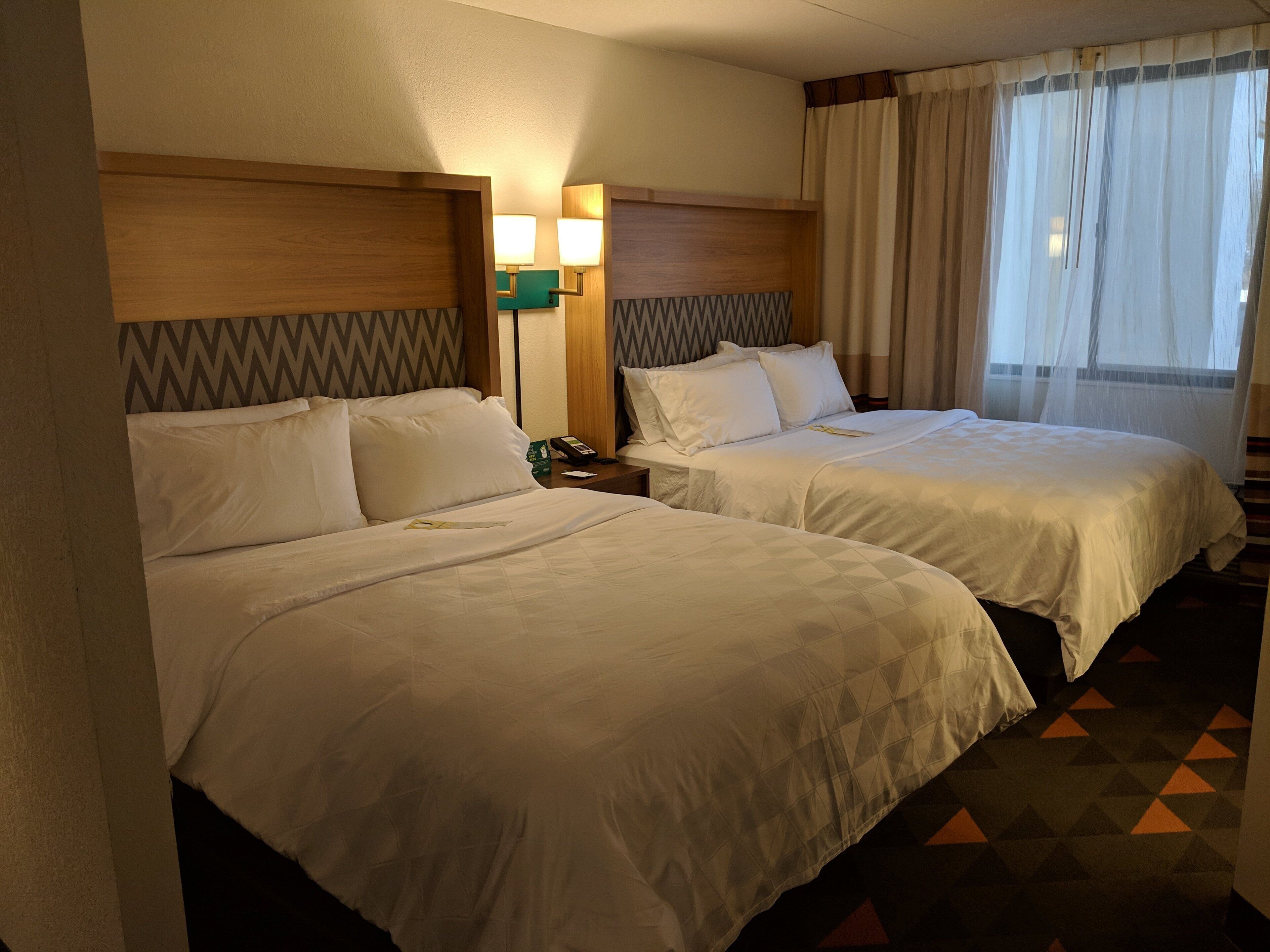 A hotel room with two beds, a nightstand with a phone, and a window with sheer curtains.