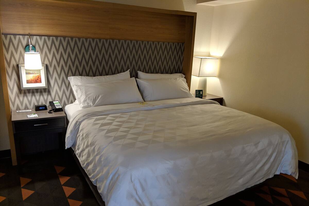 A neatly made bed with a geometric patterned bedspread, a wooden headboard, a nightstand with a lamp, and a wall clock.