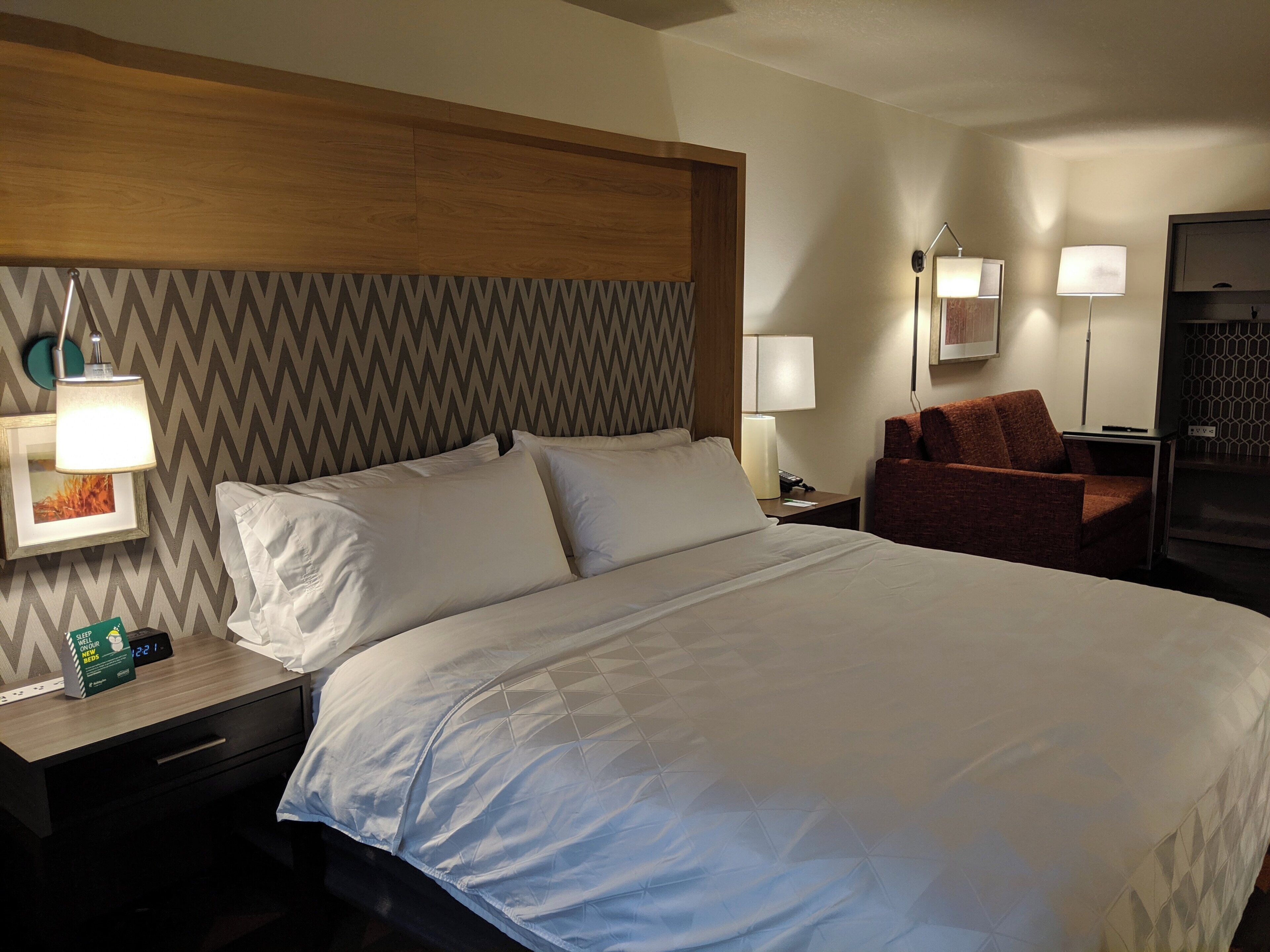 A hotel room with a large bed, two bedside tables, a nightstand, a red armchair, and a wall-mounted lamp.