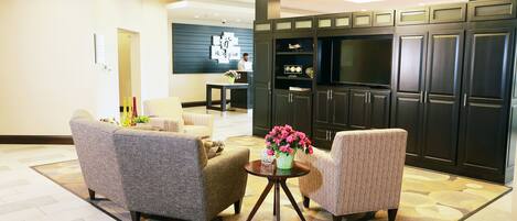A modern living room with two grey armchairs, a small coffee table with a vase of flowers, and a kitchen area with dark cabinets and a large TV.