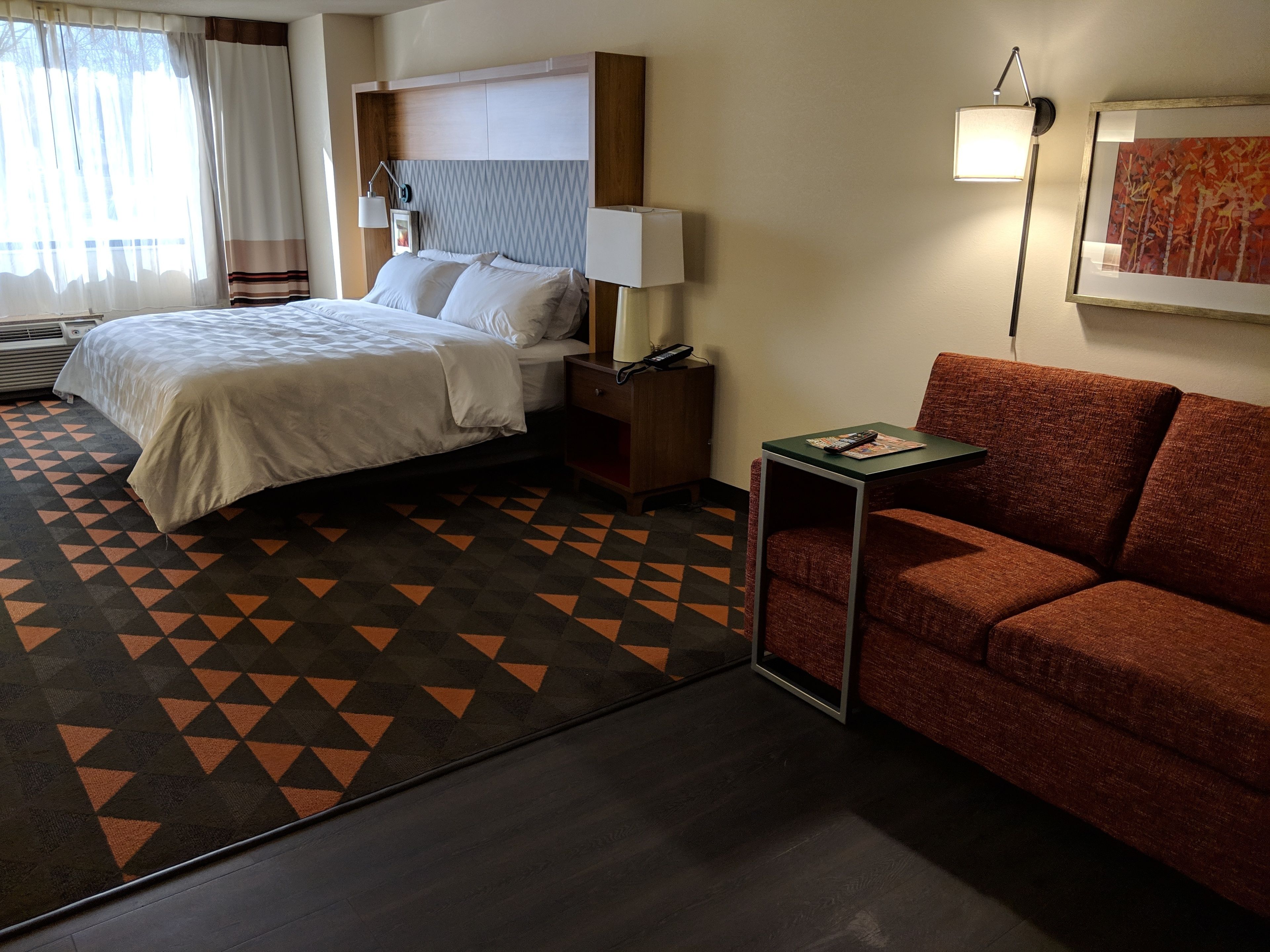 A hotel room with a bed, a nightstand, a red sofa, and a patterned rug.