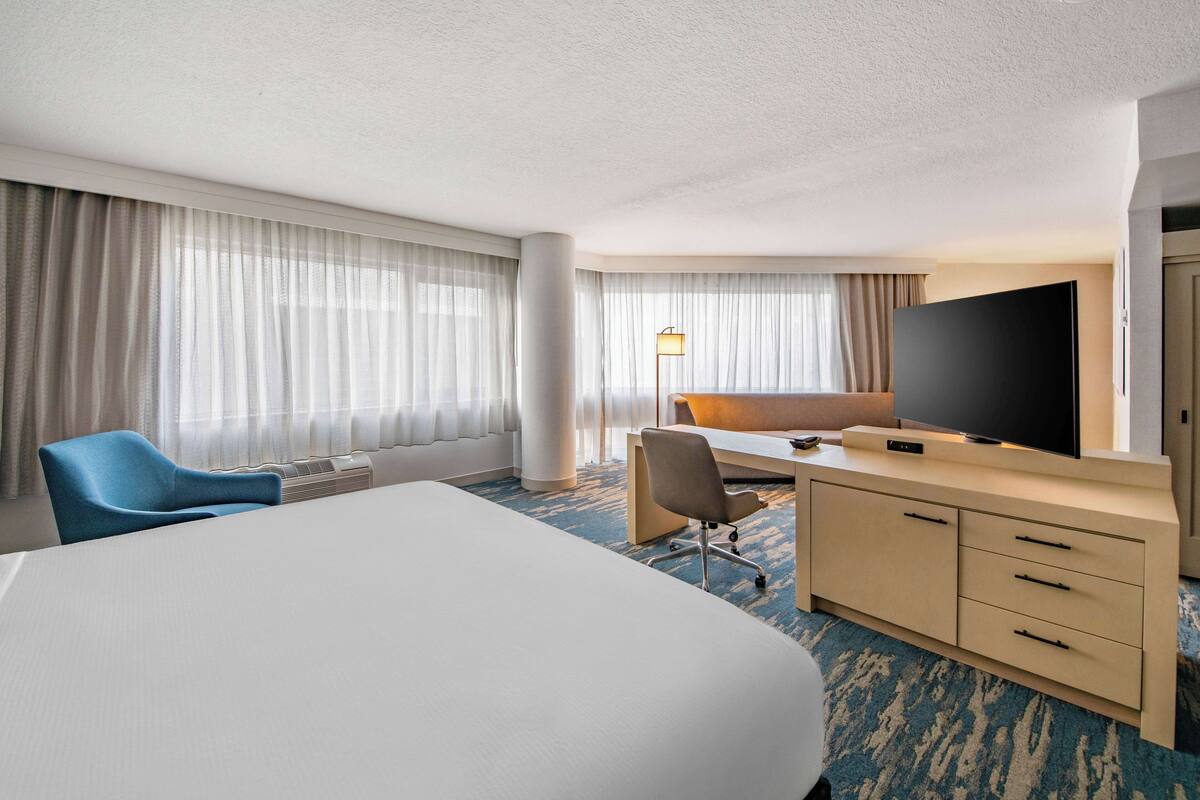 A hotel room with a large bed, a desk with a flat-screen TV, a chair, and a desk lamp.