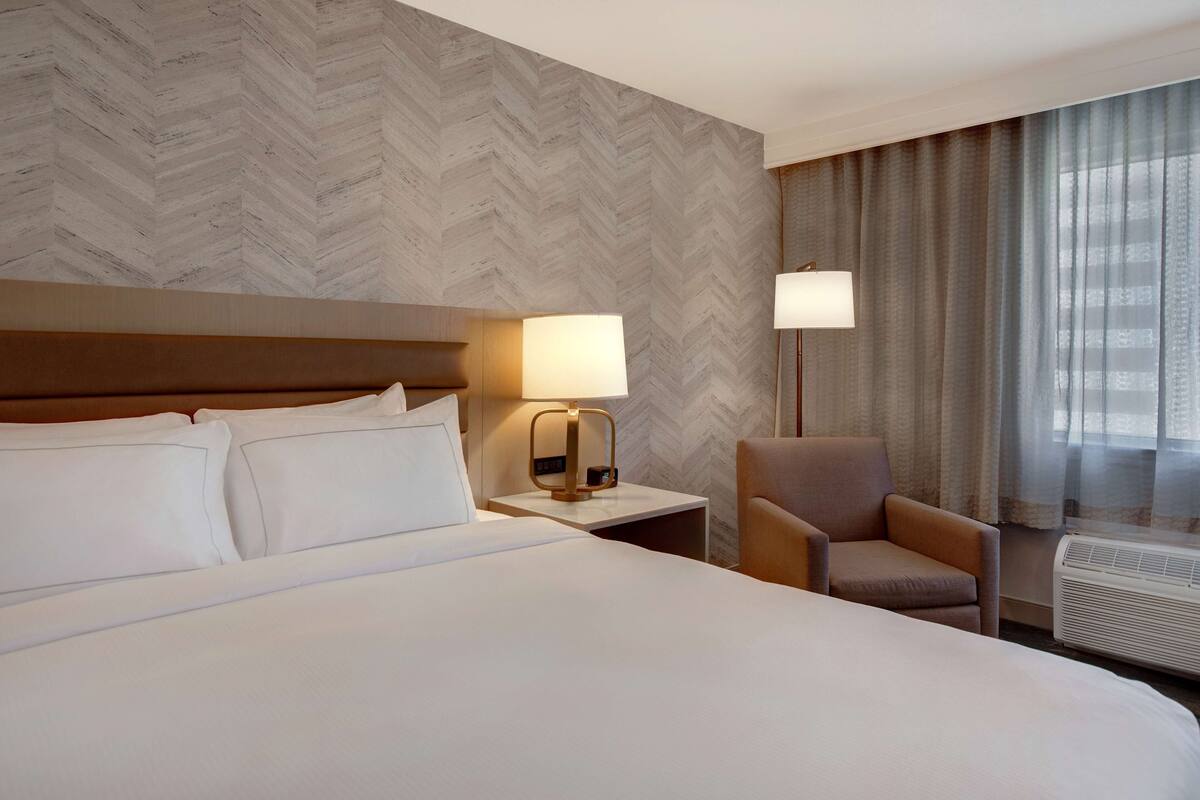 A hotel room with a large bed, a nightstand with a lamp, a chair, and a window with curtains.
