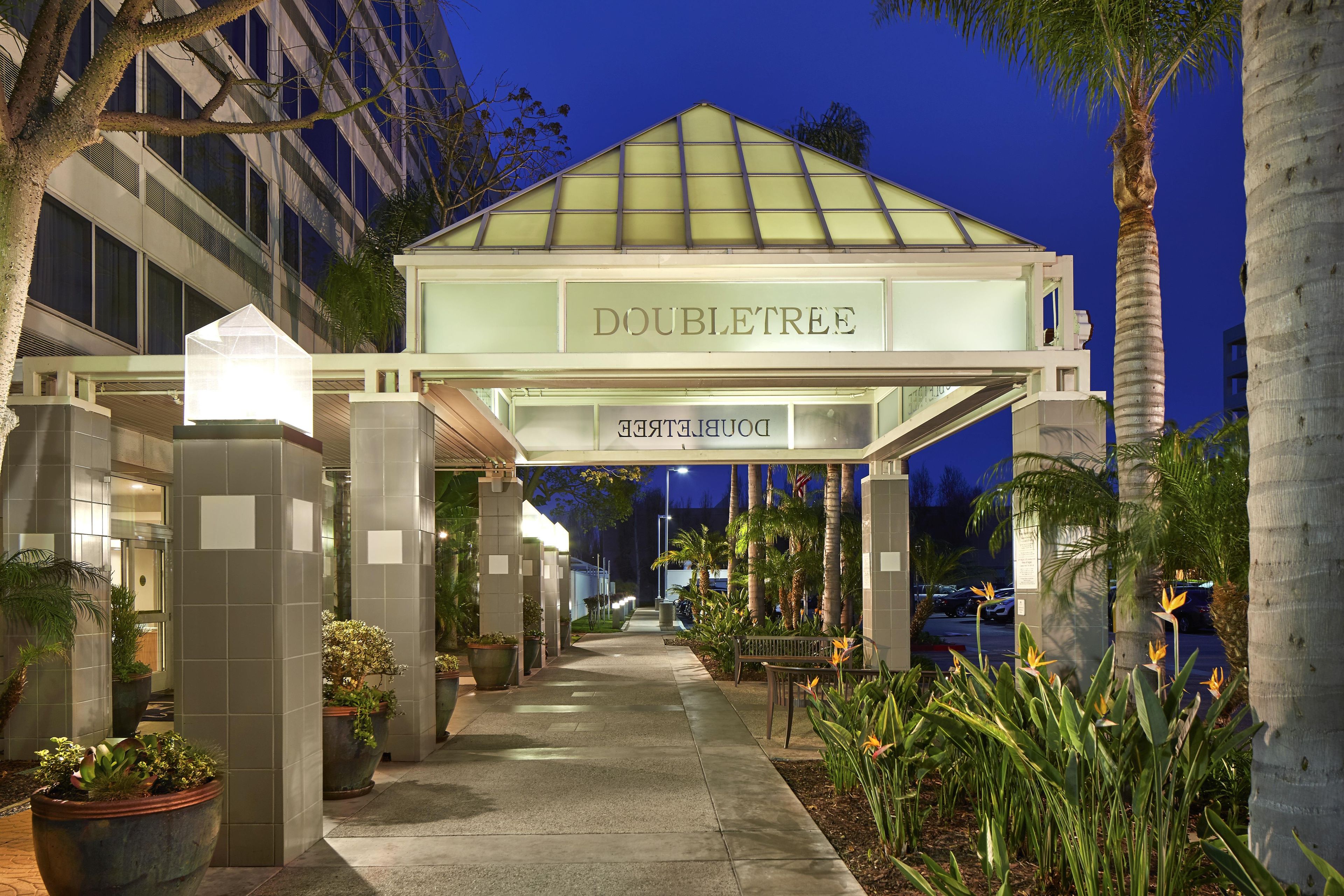 A hotel entrance with a covered walkway, palm trees, and potted plants.