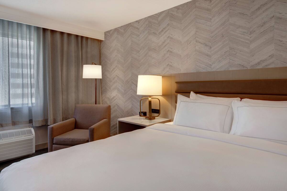 A hotel room with a large bed, a chair, a nightstand with a lamp, and a window with curtains.