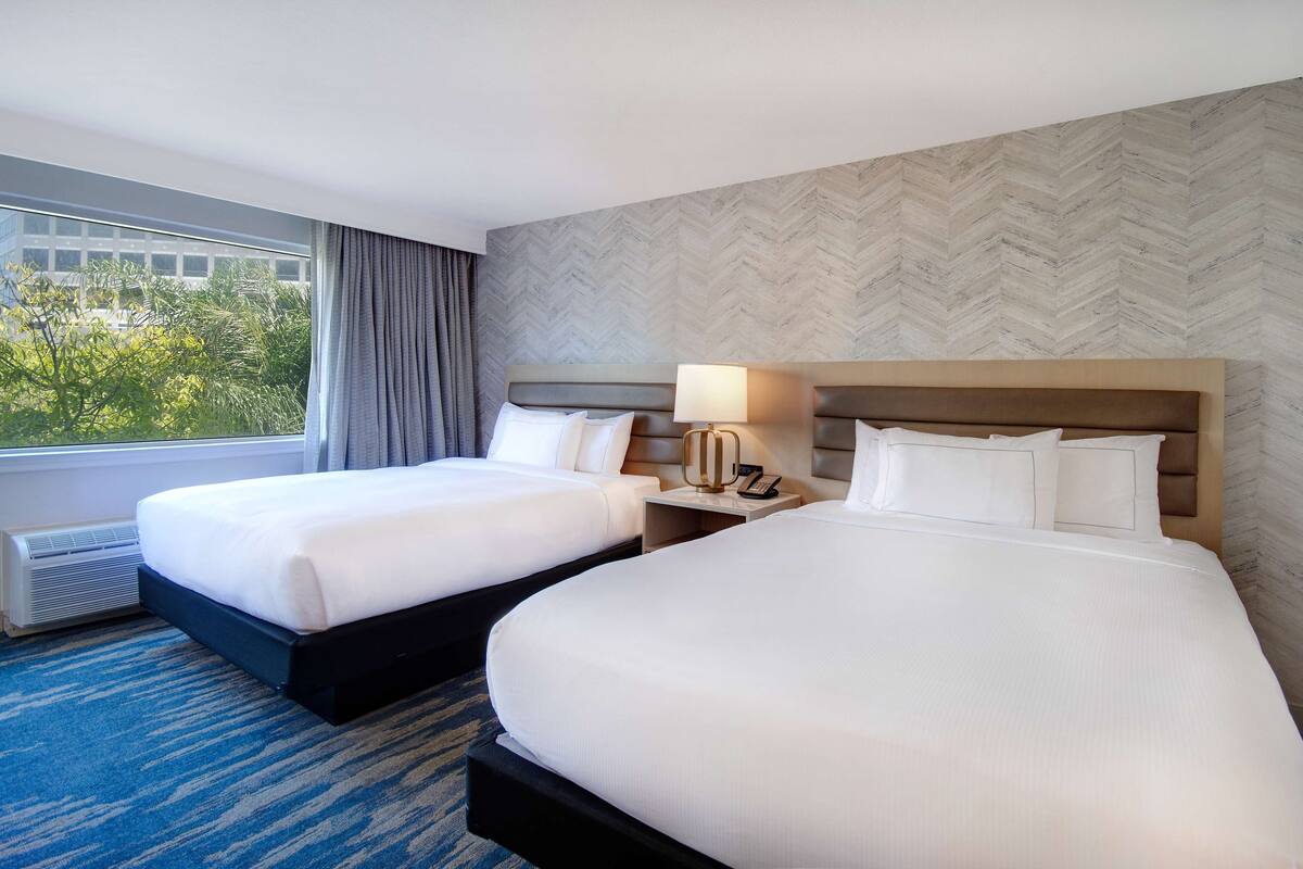 A hotel room with two beds, a large window with curtains, a wall with a herringbone pattern, and a nightstand with a lamp and phone.