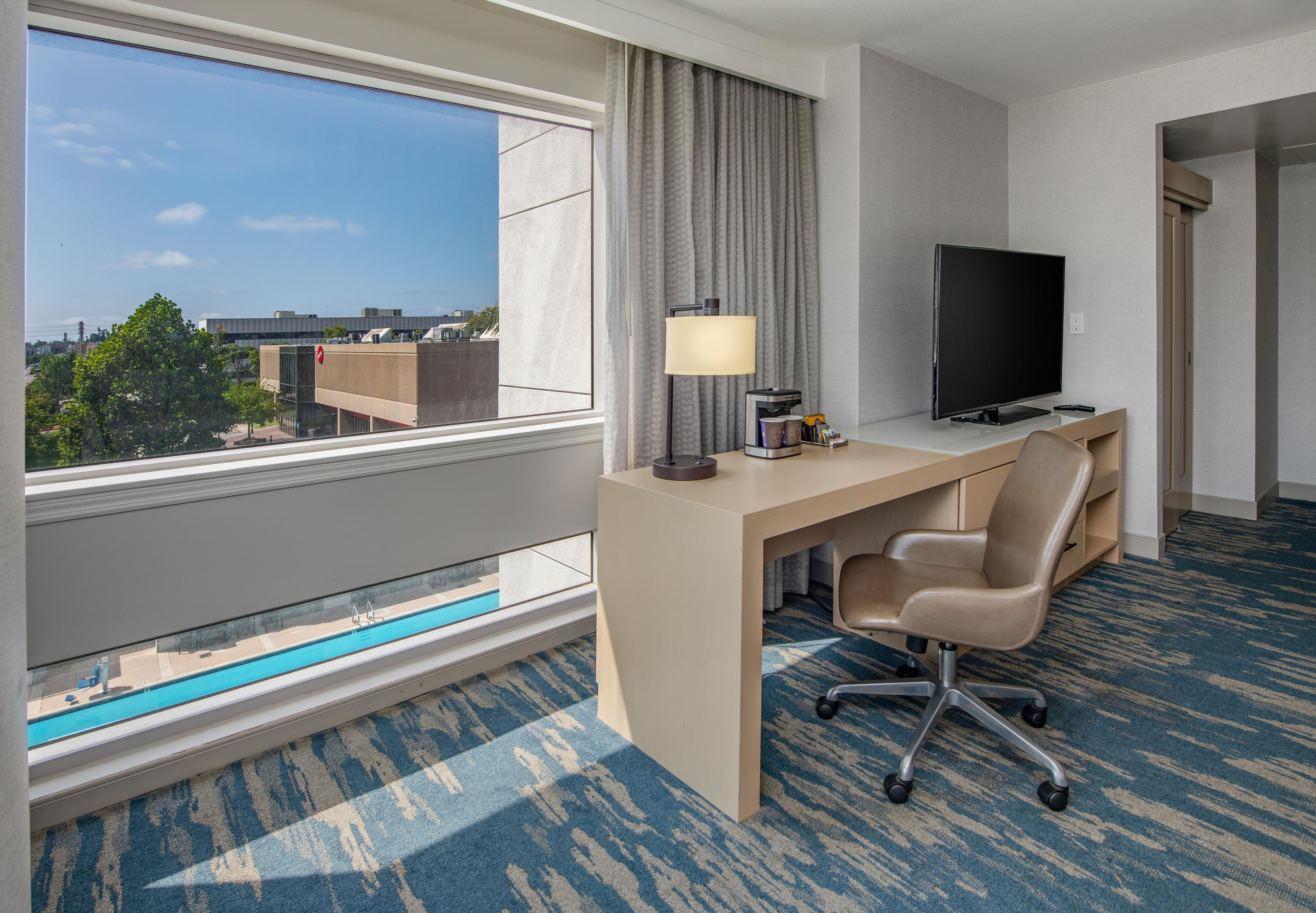Habitación de hotel con escritorio, silla, televisor y vista panorámica de la ciudad a través de la ventana.